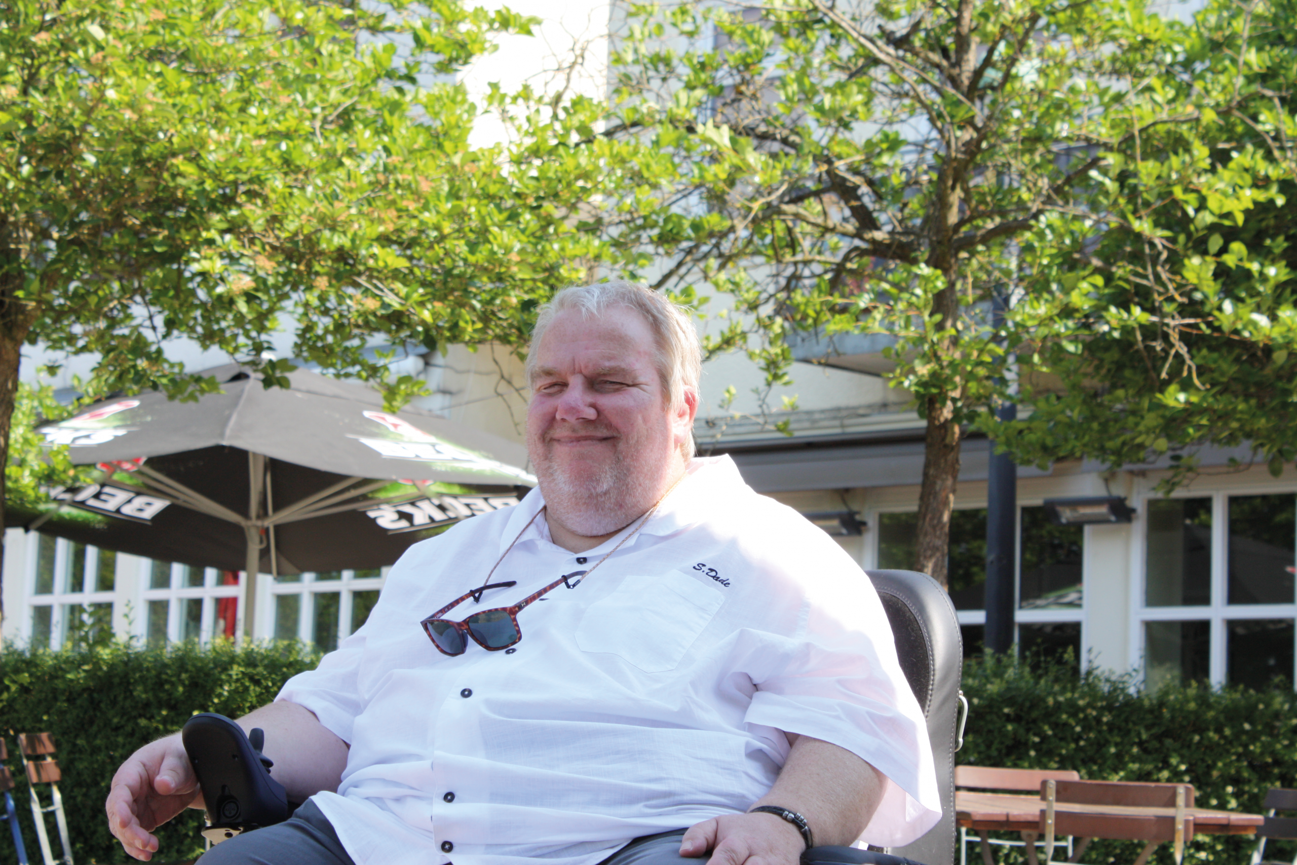 Ein lächelnder Mann mit kurzen, grauen Haaren sitzt in einem Elektrorollstuhl. Er trägt ein weißes Hemd und Sonnenbrillen. Im Hintergrund sind Bäume und ein Sonnenschirm mit dem Schriftzug „Bier“ zu sehen.