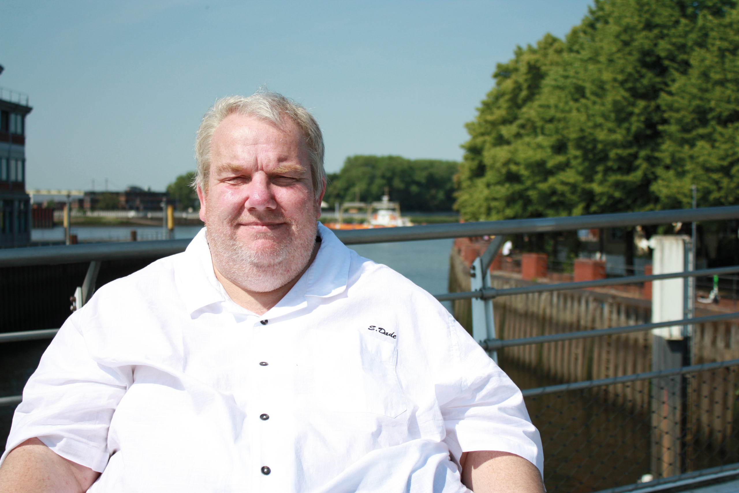 Ein lächelnder Mann mit kurzem, grauem Haar sitzt vor einem Gewässer. Er trägt ein weißes Hemd und blickt entspannt in die Kamera. Im Hintergrund sind Bäume und ein Boot zu sehen, sowie eine sonnige, klare Himmel.