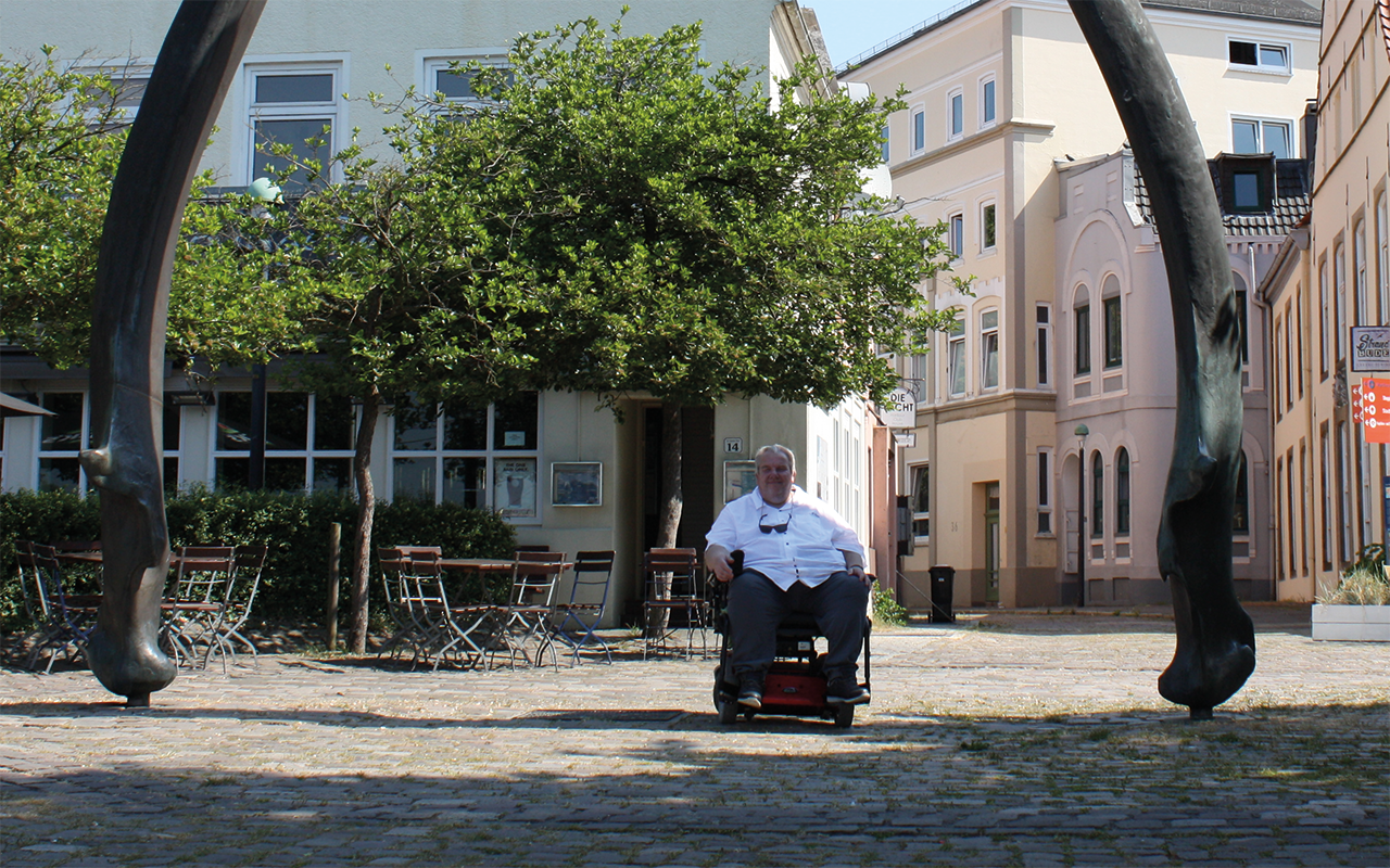 Ein Mann in einem weißen Hemd sitzt lächelnd in einem Rollstuhl auf einer gepflasterten Straße. Im Hintergrund sind Gebäude und Bäume zu sehen, sowie eine große Statue.